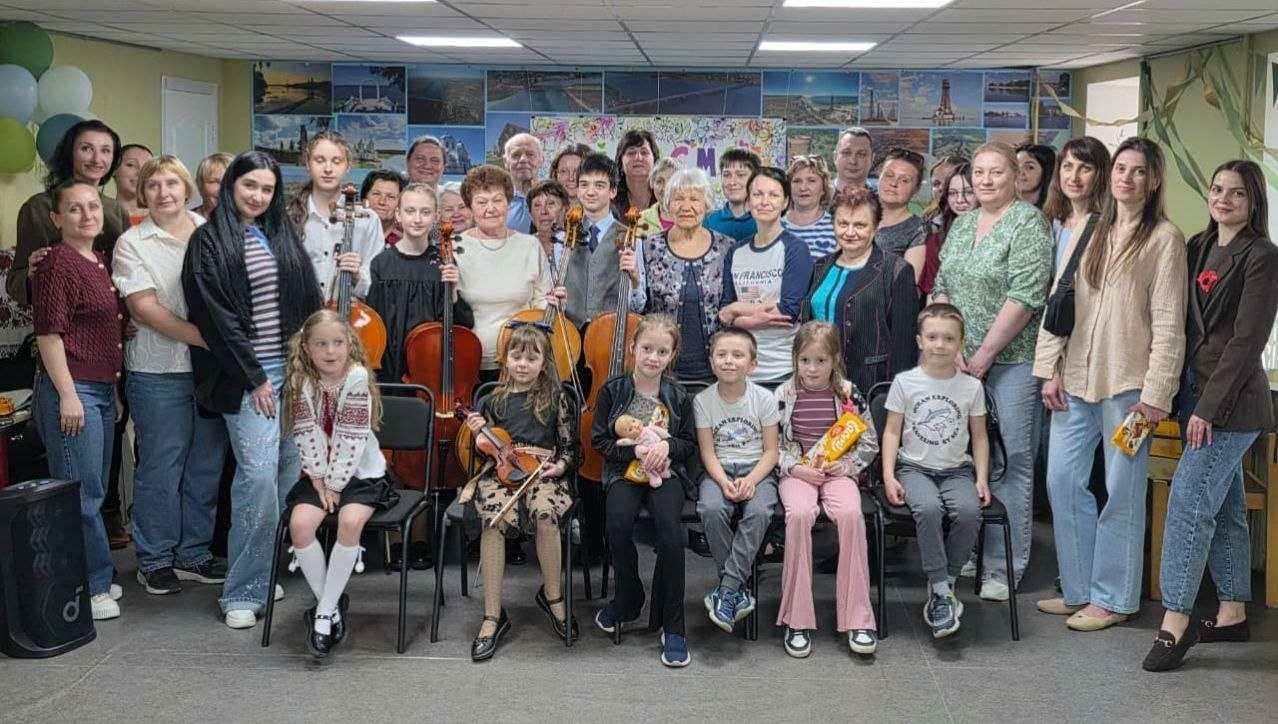 Велика група людей різного віку — дорослі та діти — позує для спільного фото у приміщенні. Серед присутніх є діти з музичними інструментами (віолончелями та скрипкою), кілька дівчаток і хлопчиків сидять на стільцях попереду, а на задньому плані стіна прикрашена численними фотографіями.