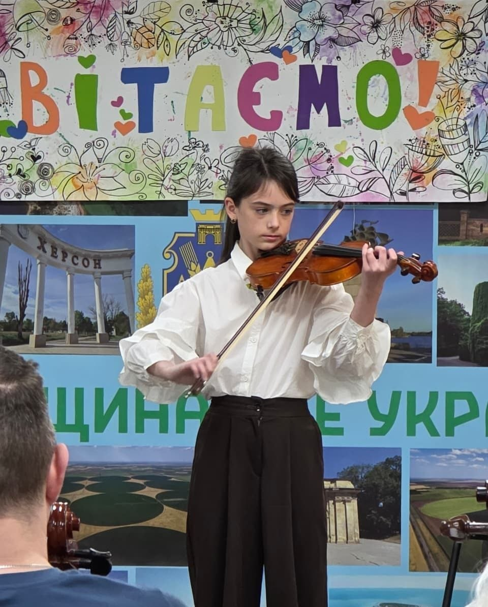 Дівчинка у білій блузі та темних штанах виступає на сцені, граючи на скрипці. На задньому плані видно частину банера з великим кольоровим написом «Вітаємо!» та фотоколаж із краєвидами.