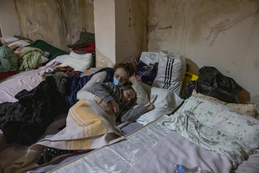 Жінка у захисній масці лежить на облаштованому на підлозі ліжку в бомбосховищі або підвалі та обіймає сплячу дитину, вкриту ковдрою. Навколо них розкладені інші матраци з постільною білизною, сумки та пакети з речами на тлі обшарпаних бетонних стін.