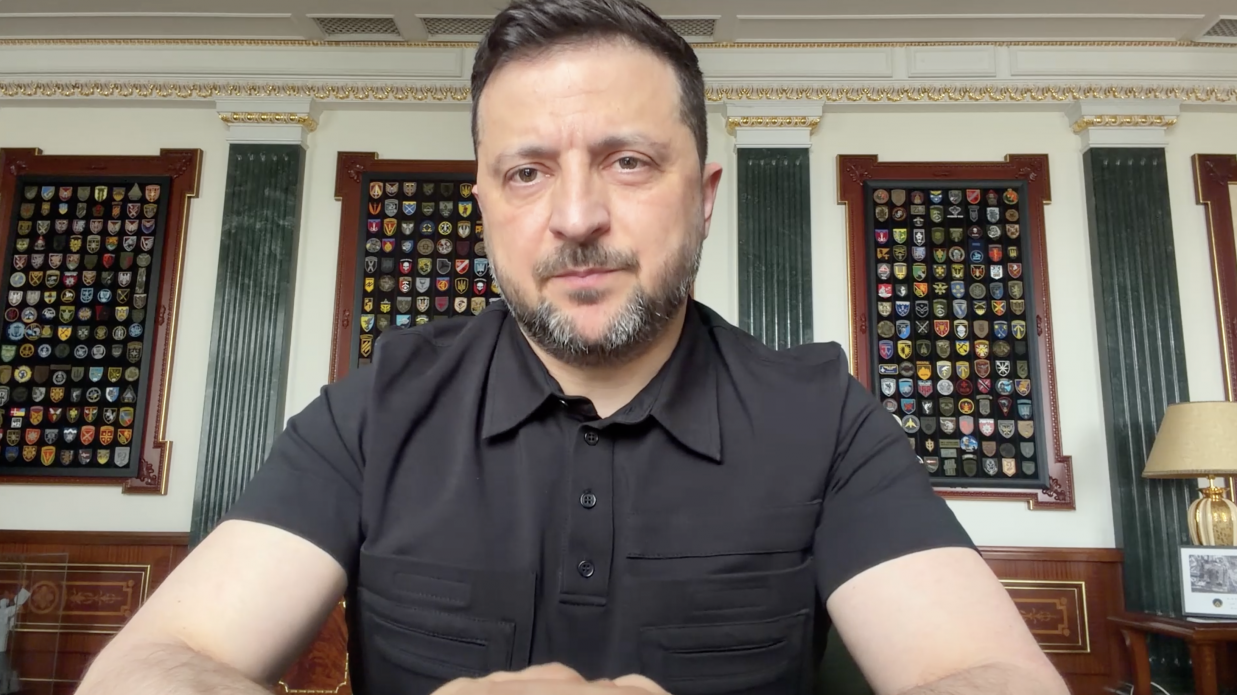 Чоловік із короткою бордою, одягнений у чорну сорочку поло, зафіксований на передньому плані великим планом. Він сидить у кабінеті, де на стінах позаду нього розвішані великі стенди з численними шевронами та військовими емблемами.