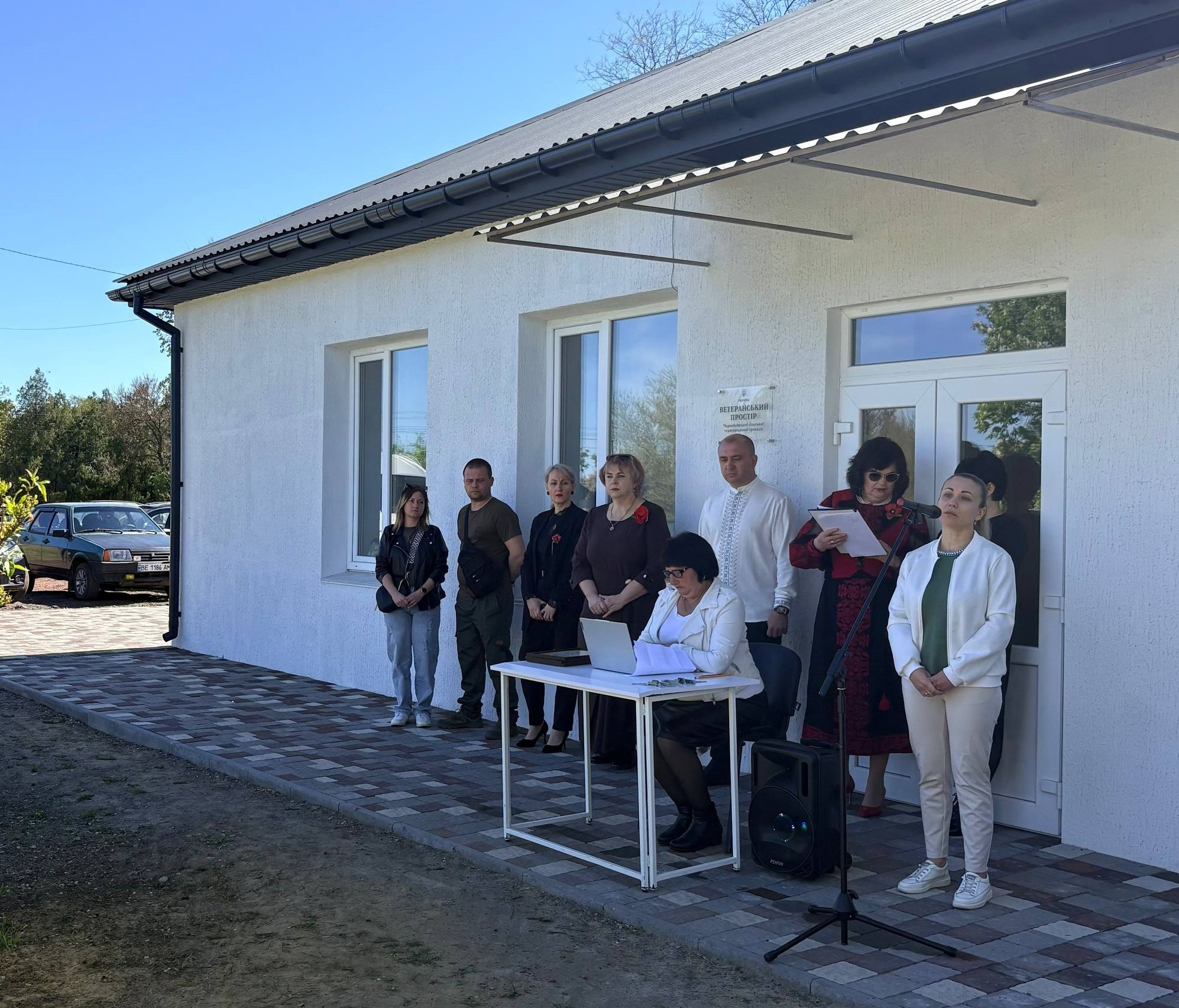 Група людей стоїть на офіційному заході біля входу до одноповерхової білої будівлі. Перед ними встановлено стіл із ноутбуком та мікрофон на стійці, а одна з жінок зачитує текст із аркуша.