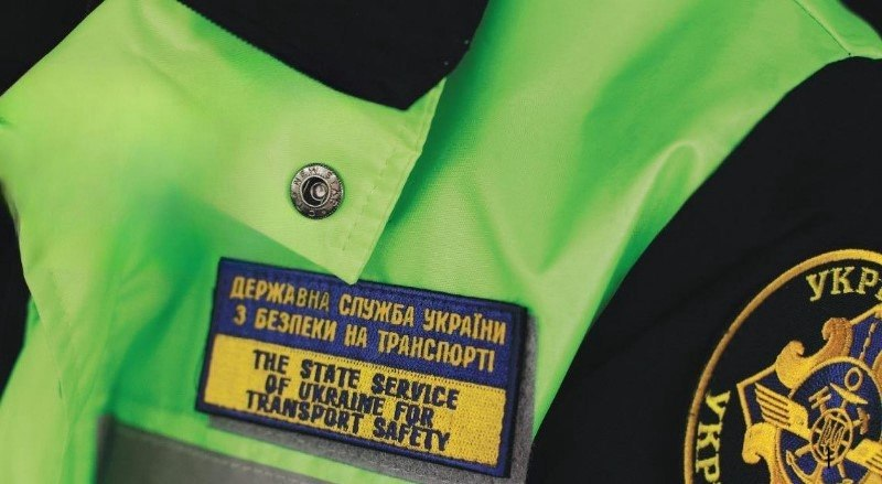 Формена куртка працівника Державної служби України з безпеки на транспорті. На яскраво-зеленому жилеті закріплено нашивку з назвою відомства українською та англійською мовами. Праворуч видно частину круглого шеврона.
