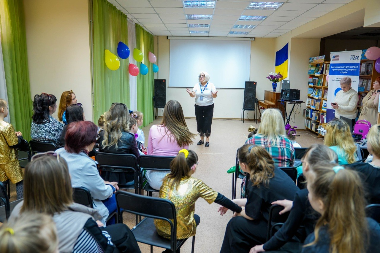 Жінка-ведуча виступає перед аудиторією на заході в херсонському центрі психічного здоров’я та психосоціальної підтримки. У залі на стільцях сидять дорослі та діти, приміщення прикрашене повітряними кульками, державним прапором України та інформаційним банером Project HOPE.