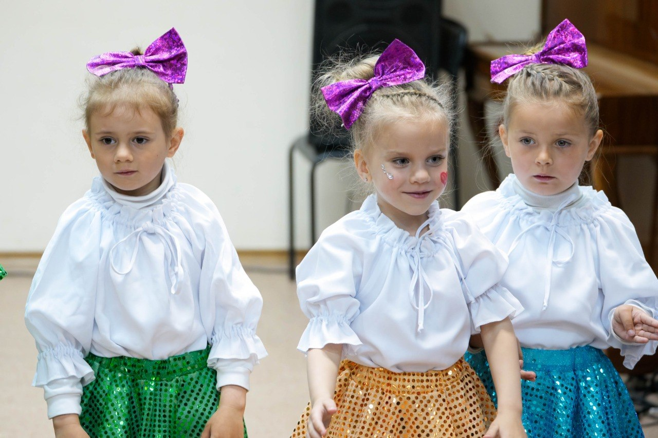 Три дівчинки у сценічних костюмах беруть участь у заході в херсонському центрі психічного здоров’я та психосоціальної підтримки. Вони одягнені в однакові білі блузи, пишні блискучі спідниці різних кольорів (зелена, жовта, блакитна) та великі фіолетові банти на голові.