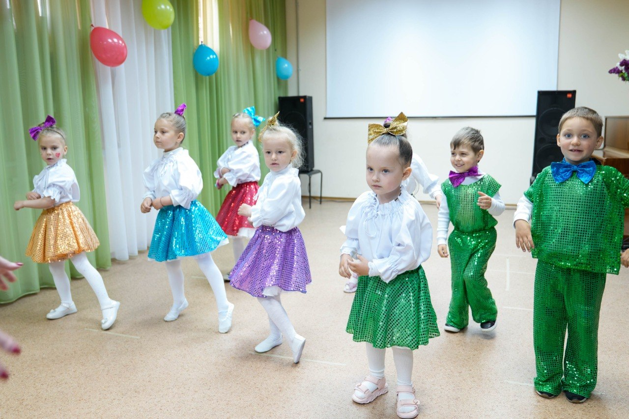 Група дітей у яскравих сценічних костюмах виступає на заході в херсонському центрі психічного здоров’я та психосоціальної підтримки. Дівчатка одягнені в білі блузи та блискучі різнокольорові спідниці з бантами на голові, а хлопчики — у зелені блискучі костюми з метеликами.