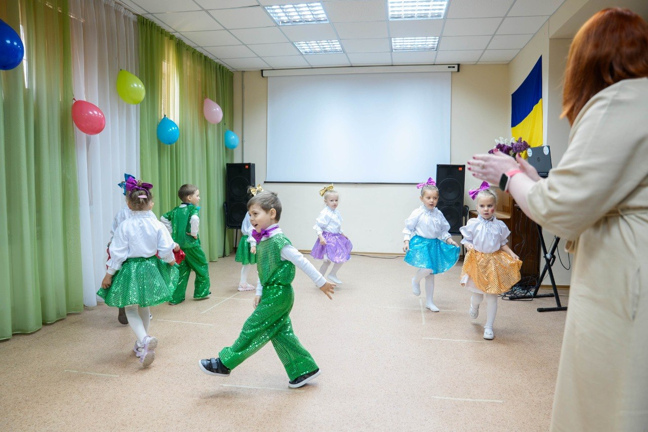 Діти у яскравих сценічних костюмах танцюють у залі під час заходу в херсонському центрі психічного здоров’я та психосоціальної підтримки. Праворуч на передньому плані стоїть жінка, яка підтримує виступ оплесками.