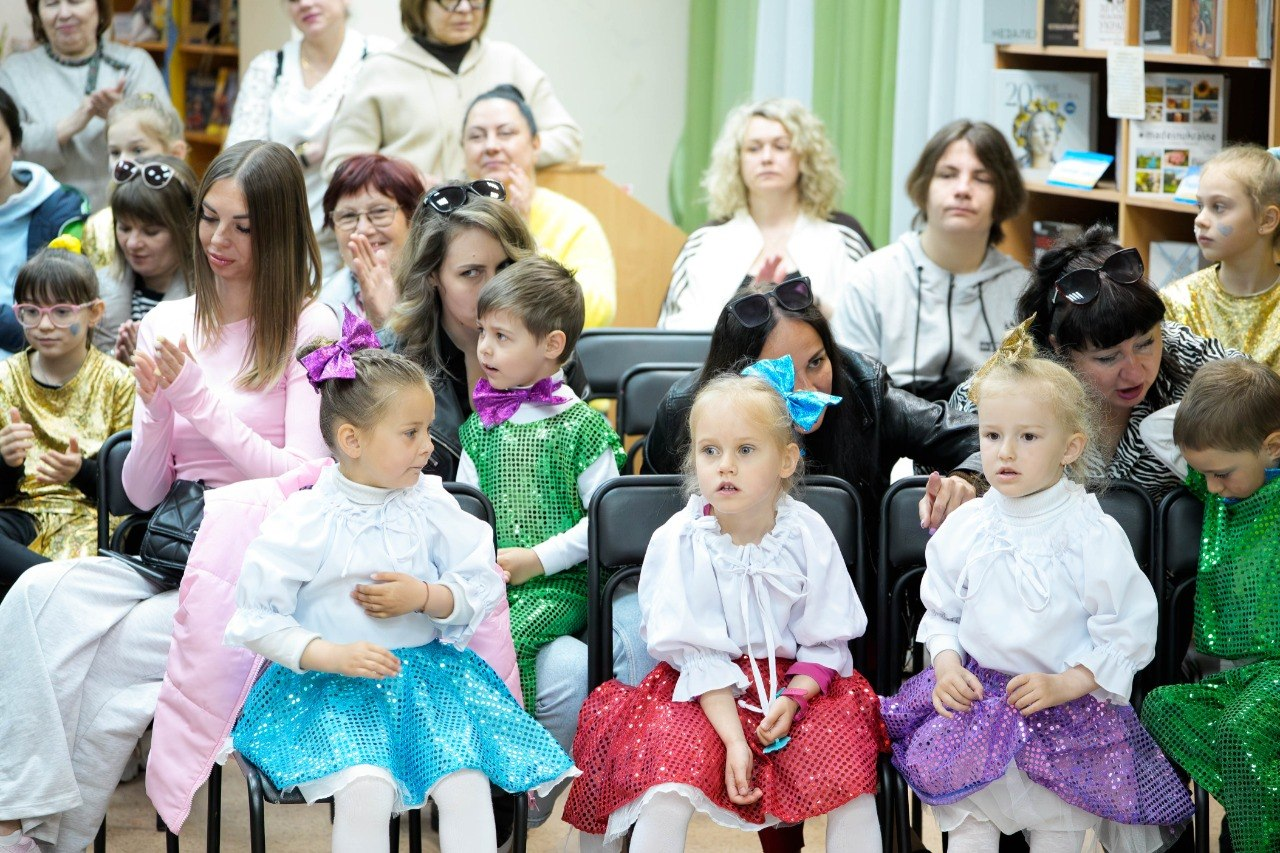 Глядачі та маленькі учасники в яскравих сценічних костюмах сидять у залі під час заходу в херсонському центрі психічного здоров’я та психосоціальної підтримки. Деякі дорослі аплодують, підтримуючи виступи дітей.