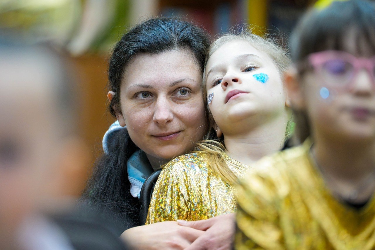 Жінка обіймає дівчинку в блискучому золотистому костюмі з малюнком на щоці під час заходу в херсонському центрі психічного здоров’я та психосоціальної підтримки. На передньому та задньому планах видно розмиті силуети інших присутніх.