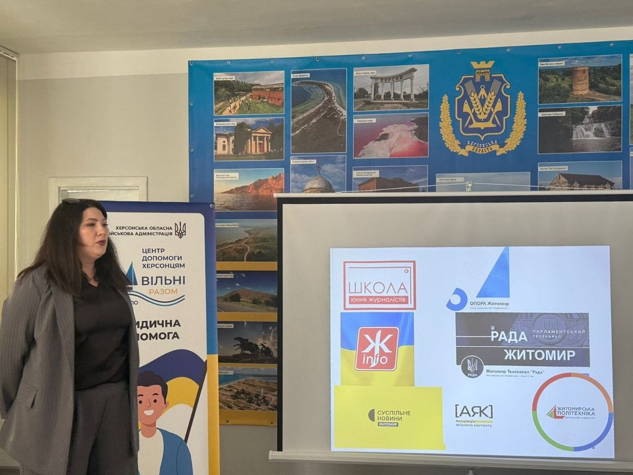Жінка виступає біля екрана зі слайдом партнерських організацій та банера Центру допомоги херсонцям «Вільні разом». На стіні позаду розміщено великий стенд із краєвидами та гербом Житомирщини.