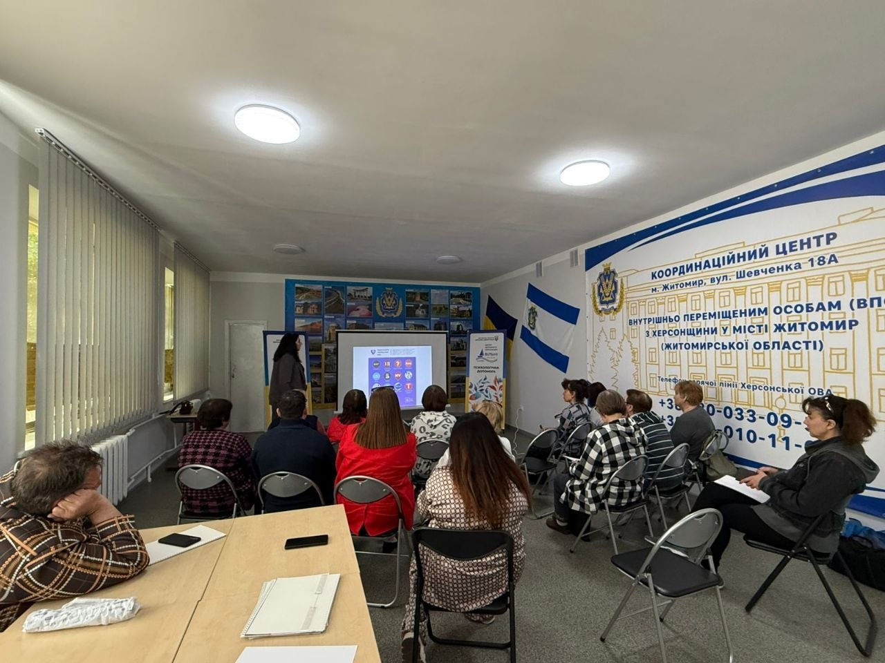 Жінка проводить презентацію біля проєкційного екрана в залі Координаційного центру для ВПО з Херсонщини у місті Житомир. Слухачі сидять на стільцях, а на стінах розміщено прапор України та великі інформаційні банери закладу.