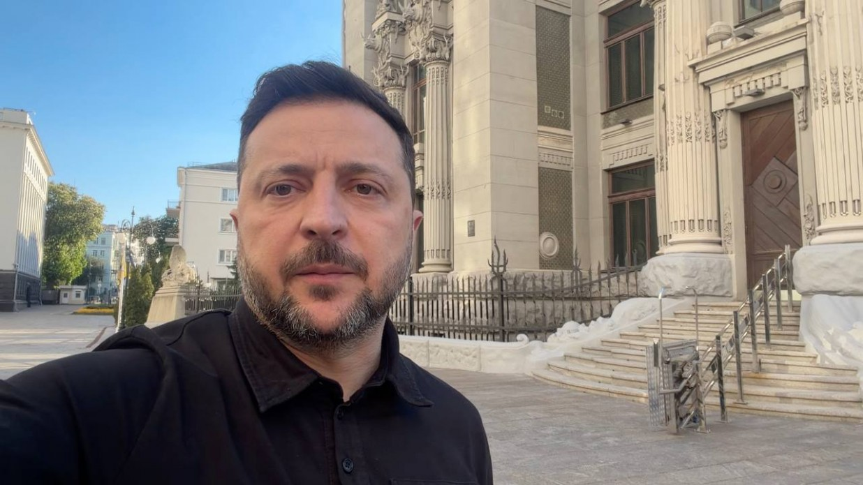 Чоловік у чорній сорочці робить селфі-знімок на вулиці на тлі великої світлої будівлі з масивними колонами, ліпниною та сходами. Ліворуч на задньому плані видно міську вулицю під ясним небом.