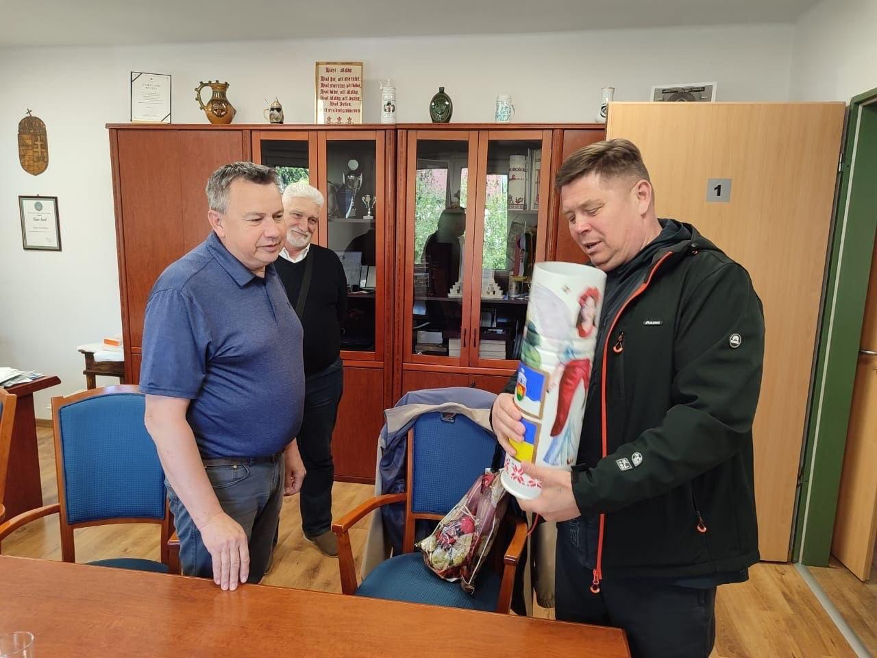 Три чоловіки перебувають у кабінеті з дерев'яними меблями. Один із них тримає в руках великий розмальований тубус із зображеннями та патріотичною символікою, а інші двоє уважно його розглядають. На задньому плані видно сервант із кубками та сувенірами, а також грамоти на стінах.