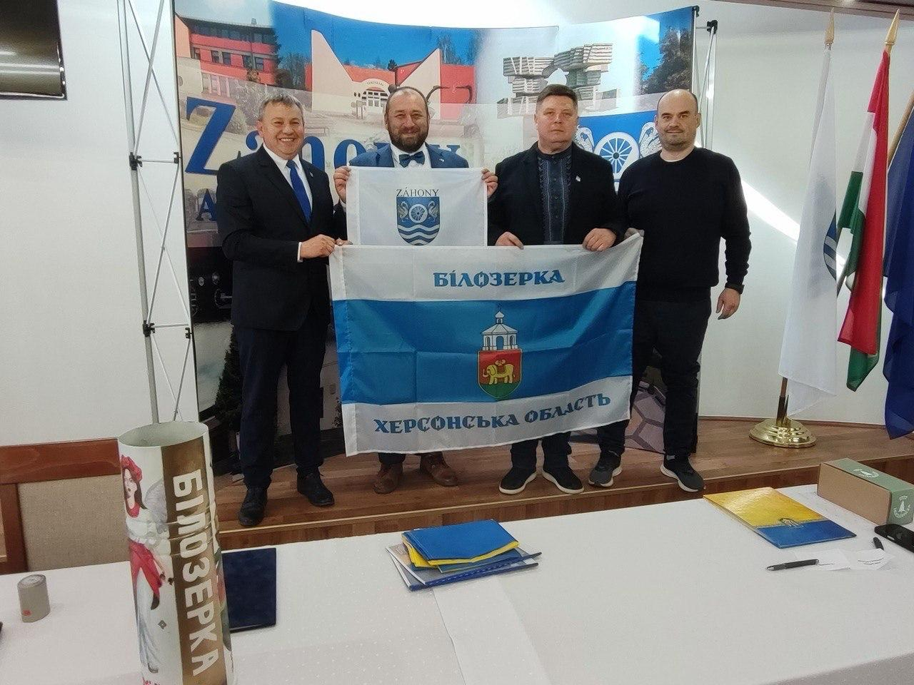 Чотири чоловіки стоять у залі та тримають два прапори: менший — білий із гербом міста Захонь, та більший — синьо-білий прапор із написом «Білозерка Херсонська область». На тлі видно інформаційний банер та державні прапори, а на передньому плані — стіл із документами та тубусом.