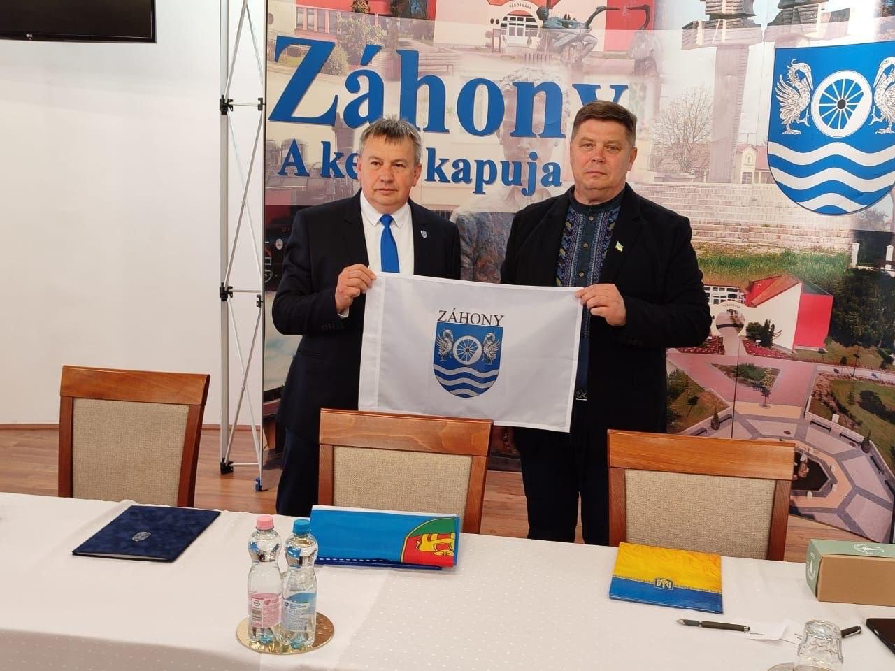 Двоє чоловіків у костюмах стоять у конференційній залі за білим столом і тримають білий прапор із гербом міста Захонь. На тлі розміщено великий банер із написами та символікою. На столі лежать папки, стоїть вода та інше приладдя.