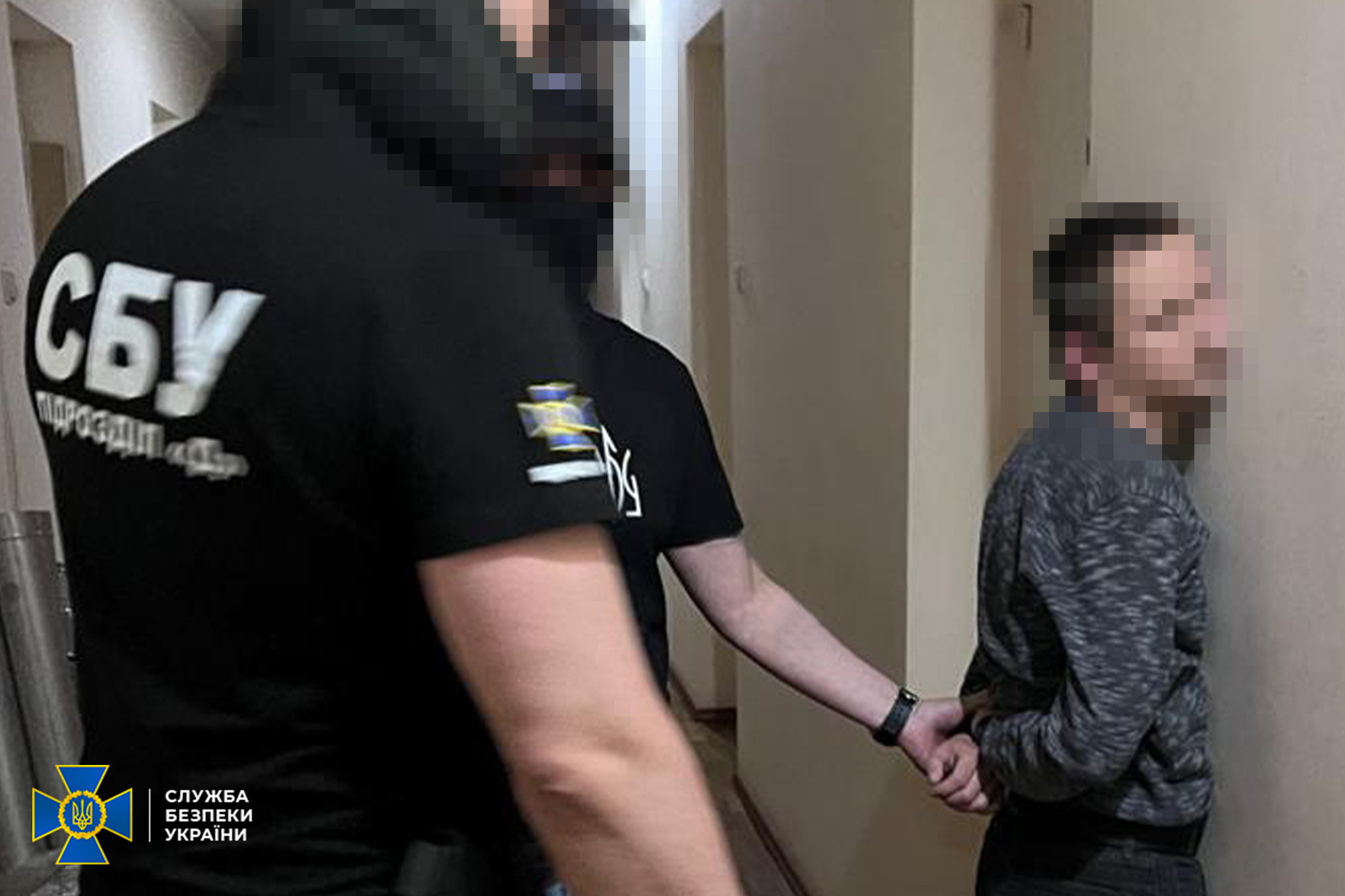 Співробітники СБУ затримують чоловіка в коридорі. Затриманий стоїть обличчям до стіни з руками за спиною, а правоохоронець у чорній футболці з написом «СБУ» утримує його. Обличчя людей розмиті. У нижньому кутку розміщено логотип Служби безпеки України.