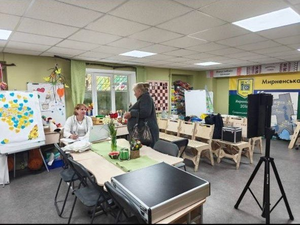 Жінка за робочим столом консультує відвідувачку у світлому приміщенні. В кімнаті розташовані стенди з патріотичною символікою, квіти, сучасне обладнання та дерев'яні меблі. На задньому плані видно банер Мирненської громади.