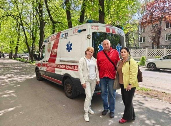 Три людини стоять поруч із автомобілем швидкої допомоги на узбіччі вулиці. На білому мікроавтобусі видно напис литовською мовою «Greitoji medicinos pagalba», червоні смуги та номери екстрених служб.
