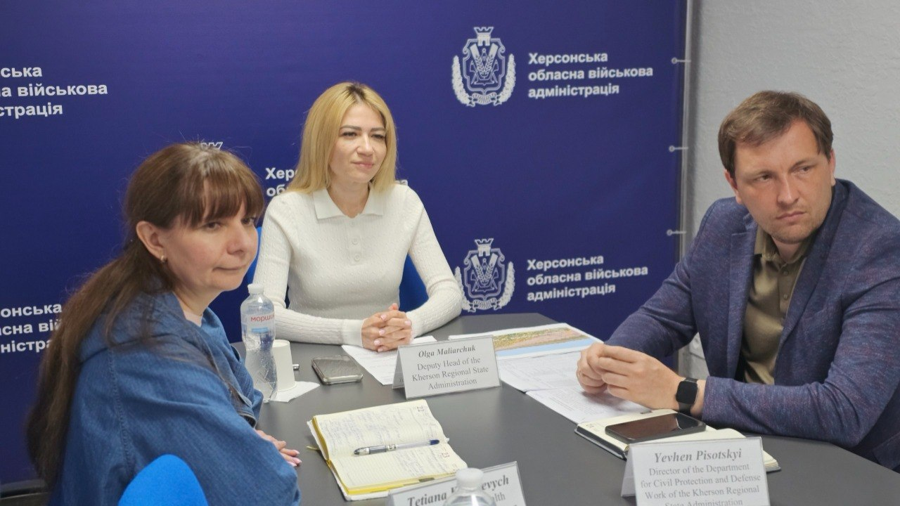 Троє людей — дві жінки та чоловік — сидять за столом під час офіційної зустрічі на фоні синього банера з державною символікою. Перед ними на столі лежать робочі документи, блокноти, мобільні телефони та іменні таблички.