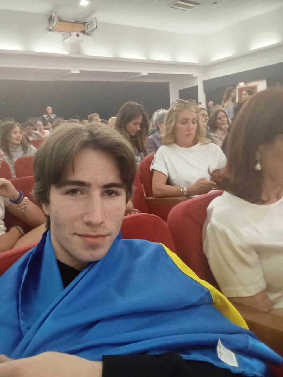 Молодий чоловік із прапором України на плечах сидить у глядацькій залі серед інших людей. На фоні видно ряди червоних крісел та частину сцени в приміщенні з яскравим освітленням.