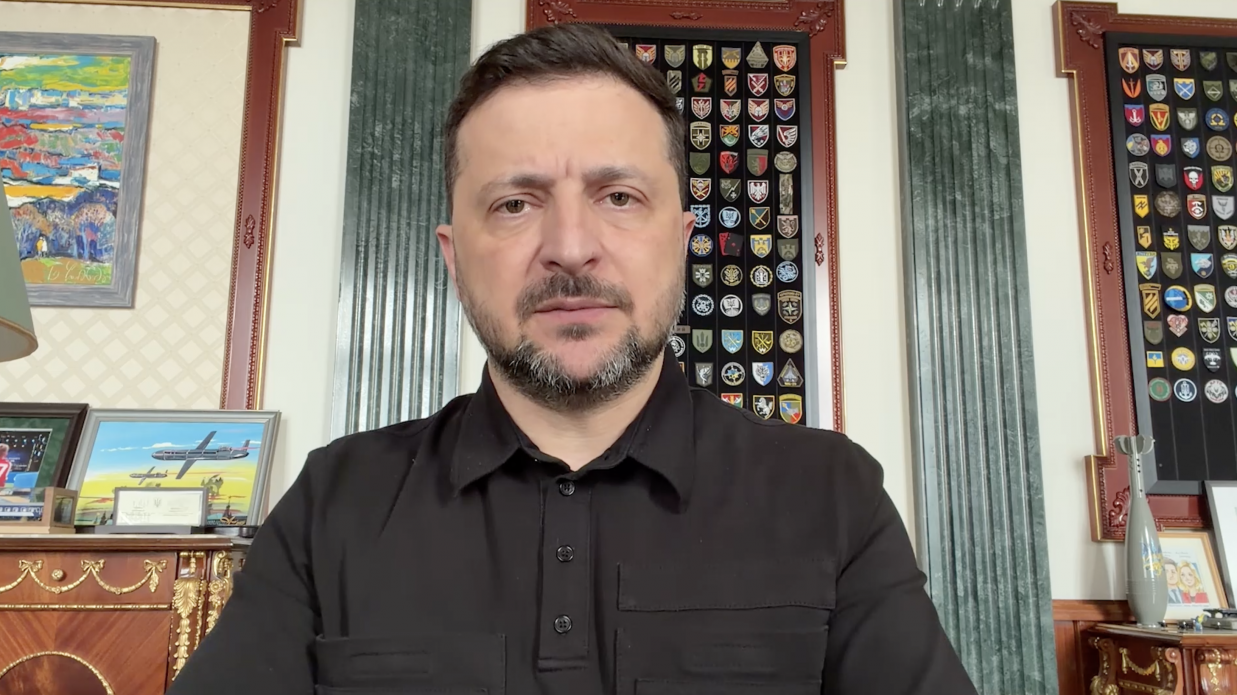 Чоловік у чорній сорочці-поло з коротким рукавом зафіксований крупним планом під час офіційного відеозвернення. На задньому плані видно інтер'єр кабінету з дерев'яними меблями, колонами, картинами та великими стендами з військовими шевронами.