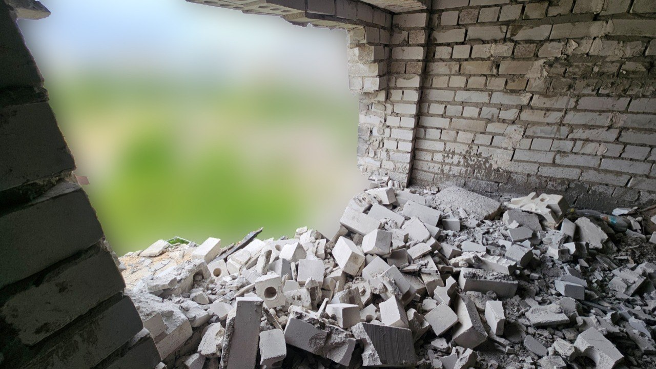 Вид зсередини пошкодженого приміщення із силікатної цегли. У стіні утворився великий пролом, під яким лежить купа уламків цегли та бетону. Краєвид за вікном штучно розмитий.