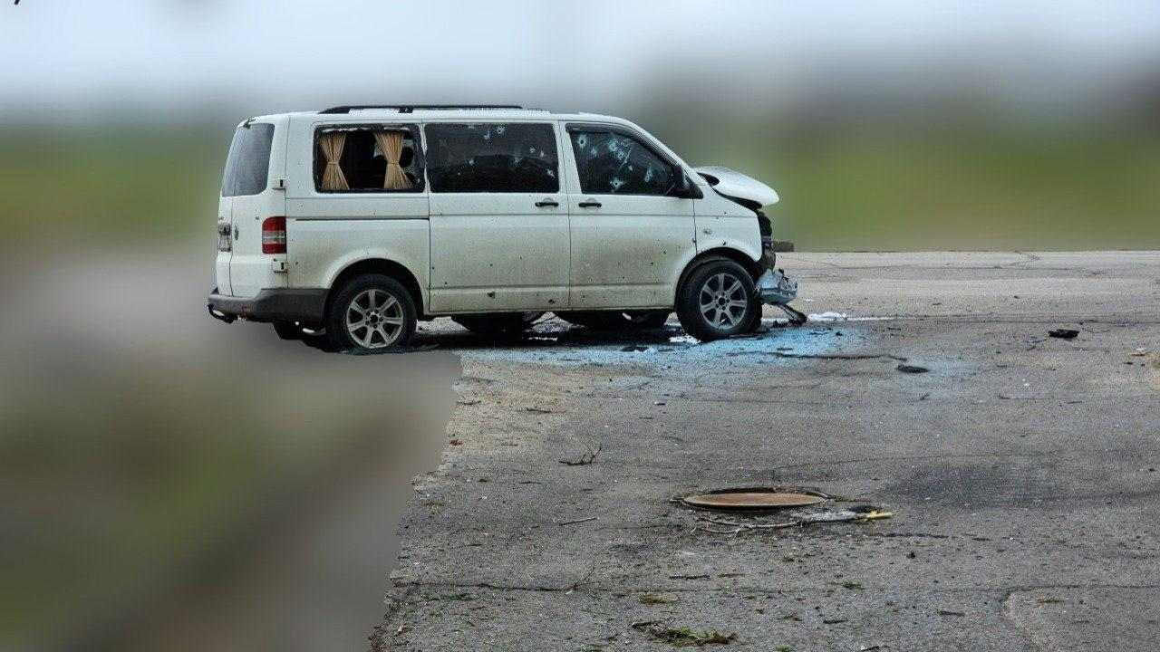 Білий пасажирський мікроавтобус із сильними пошкодженнями стоїть на узбіччі дороги. Передня частина автомобіля повністю розтрощена, лобове та бокові вікна вибиті, а на кузові видно численні отвори від уламків або куль. Навколо машини на асфальті лежить розбите скло, а фон навколо штучно розмитий.
