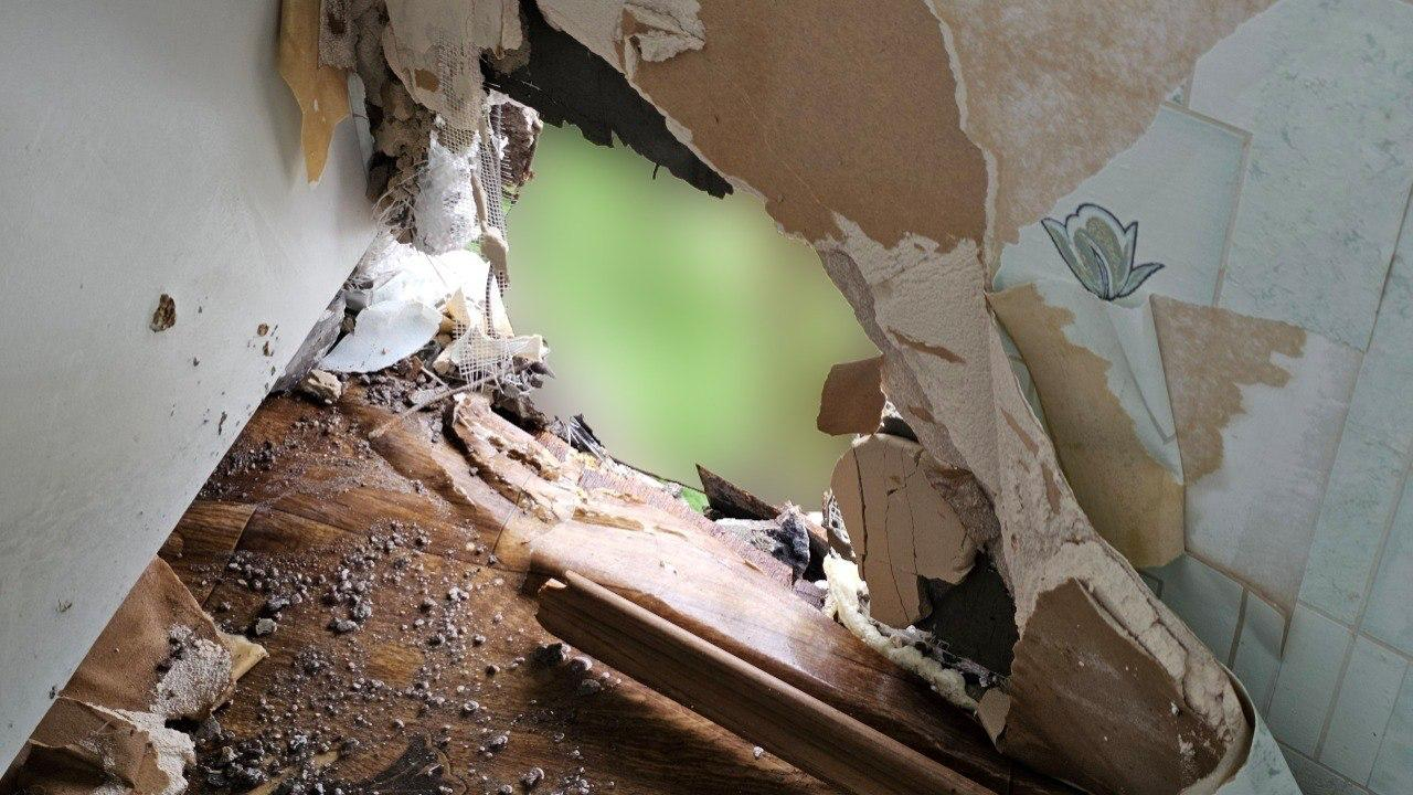 Великий наскрізний пролом у стіні кімнати, що виходить на вулицю. Навколо отвору видно обірвані шпалери, шматки штукатурки та пошкоджену кахляну плитку з візерунком, а підлога засипана будівельним сміттям і пилом. Вид на зелень за стіною штучно розмитий.
