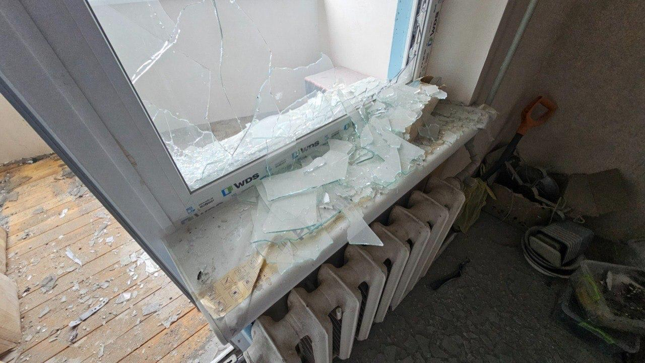 Зруйнований металопластиковий балконний блок марки WDS із повністю розбитими шибками. Великі та дрібні уламки скла рясно всипали підвіконня, радіатор опалення та підлогу в кімнаті й на балконі, де також помітно будівельний пил та речі.