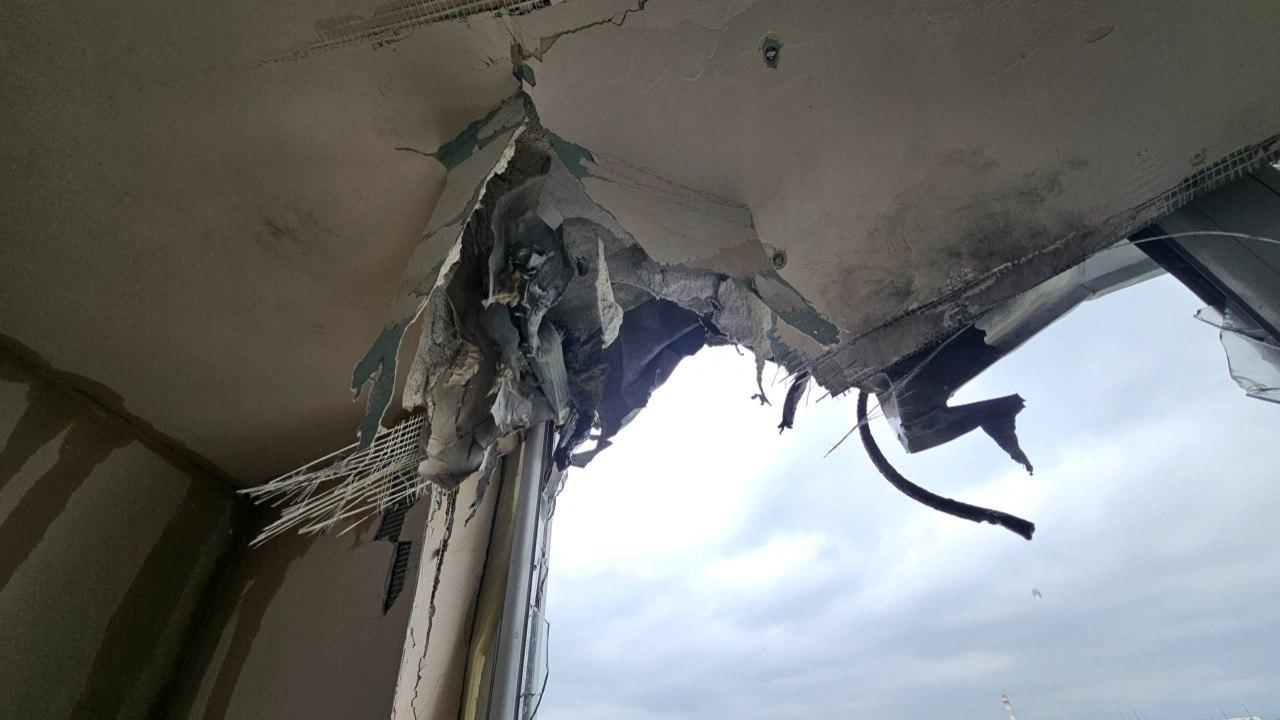 Вид знизу на сильно пошкоджений верхній кут віконного отвору та стелю в приміщенні. Бетон та штукатурка вирвані ударом, оголюючи армувальну сітку, шматки будівельних матеріалів та понівечені елементи конструкції. Через отвір видно похмуре хмарне небо.