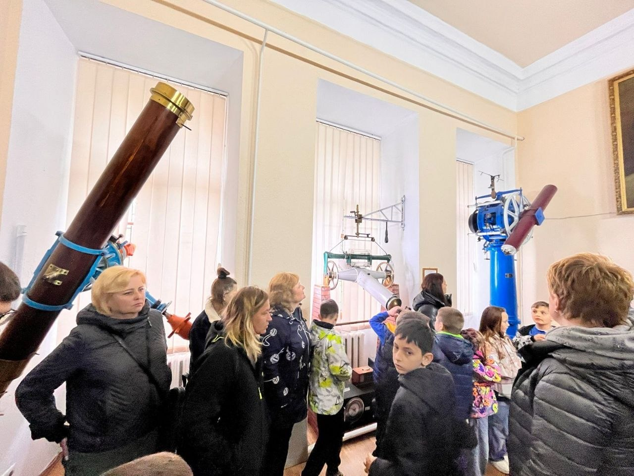 Група дітей та дорослих перебуває на екскурсії в обсерваторії. Вони розглядають старовинні астрономічні прилади та великі телескопи, що розміщені в залі біля високих вікон із жалюзі.