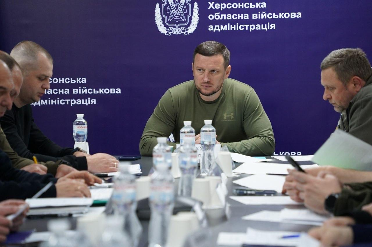 Офіційна зустріч або засідання за довгим столом. У центрі сидить чоловік у зеленій спортивній кофті, навколо нього перебувають інші учасники заходу з документами. На столі стоять пляшки з водою, а фон за людьми залишається однотонним синім з написами.