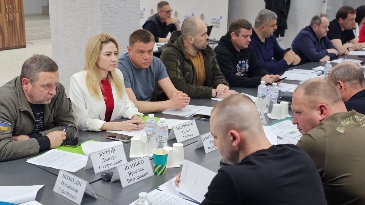Робоче засідання за великим П-подібним столом у світлому кабінеті. Учасники заходу — чоловіки та жінка в діловому й повсякденному одязі — сидять поруч із документами, паперовими табличками та пляшками води, уважно слухаючи спікера.