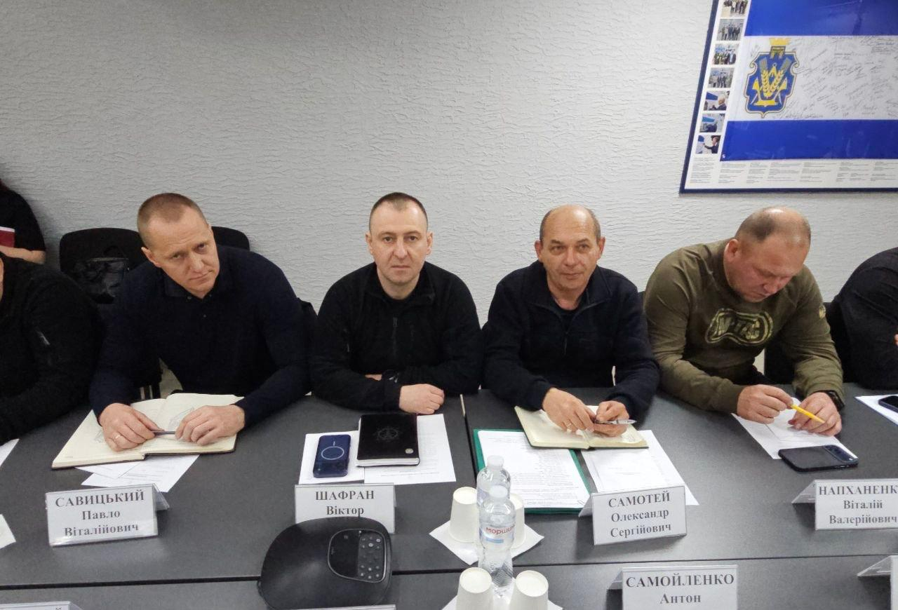 Чотири чоловіки сидять поруч за довгим столом під час засідання чи робочої наради. Перед кожним із них лежать папери, блокноти для записів та таблички. На стіні праворуч розміщено плакат із державною символікою України.