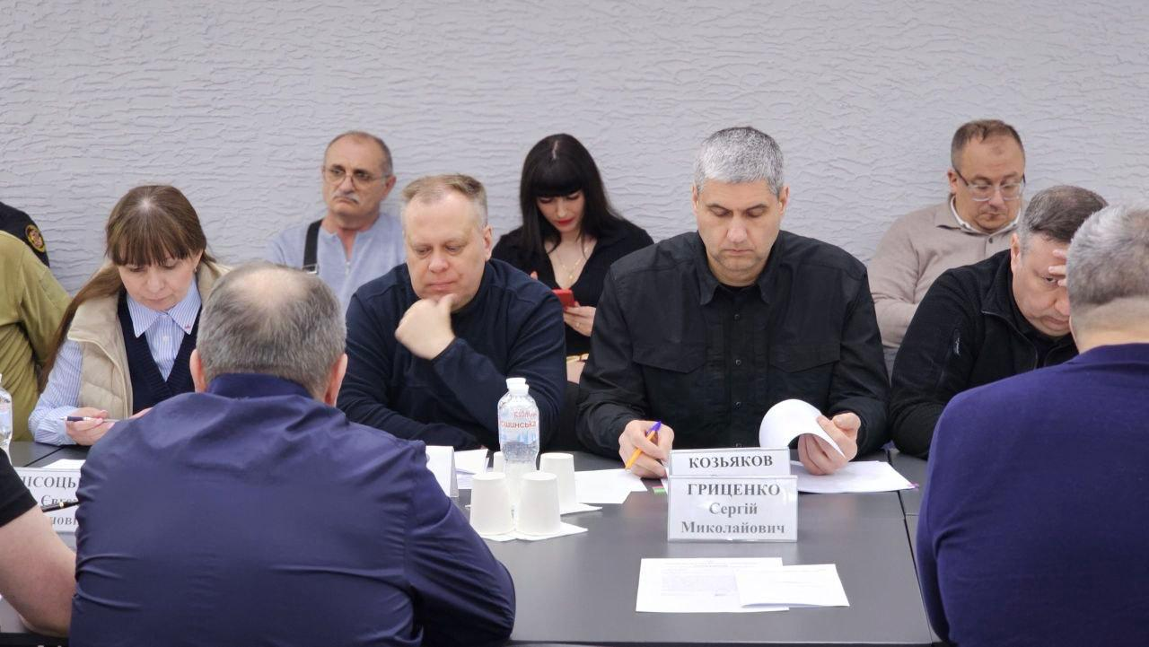Група людей бере участь у робочій нараді чи засіданні за довгим столом. Присутні чоловіки та жінки переглядають документи, роблять записи або слухають колег. На передньому плані стоять паперові стаканчики, пляшка води та інформаційні таблички.