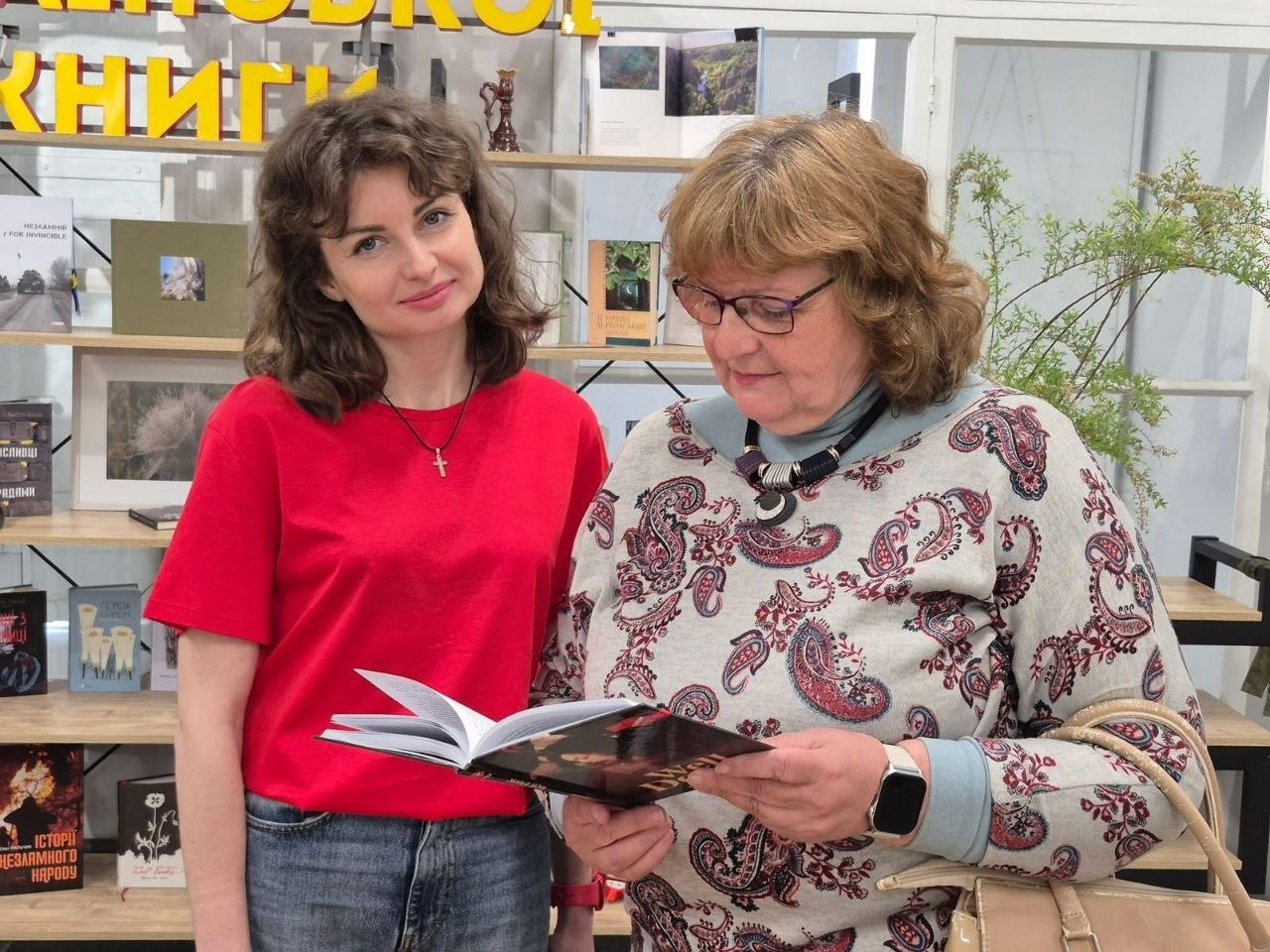 Дві жінки стоять у приміщенні на тлі стелажа з книжками. Ліворуч — молодша жінка в червоній футболці та джинсах, яка дивиться в кадр. Праворуч — жінка в окулярах та світлій кофті з візерунком «пейслі», яка тримає в руках відкриту книгу й читає її.