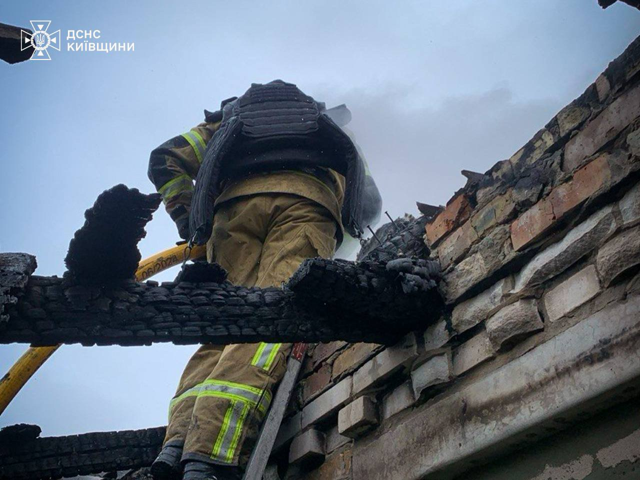 Рятувальник у захисному спорядженні стоїть на драбині біля зруйнованого даху цегляного будинку та гасить пожежу за допомогою шланга. Навколо видно обвуглені дерев'яні балки, попіл та задимлене небо. У верхньому лівому кутку розміщено логотип ДСНС Київщини.