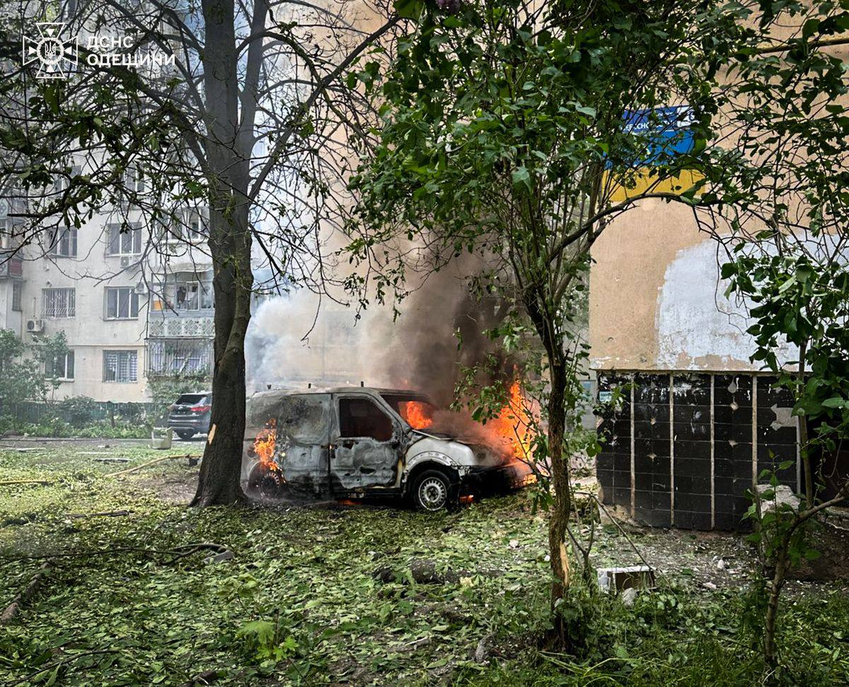 Легковий фургон повністю охоплений вогнем біля стіни багатоповерхового будинку, від машини підіймається густий чорний дим. Навколо розкидане зрізане вибухом зелене листя та гілки дерев, а в лівому верхньому кутку розміщено логотип ДСНС Одещини.