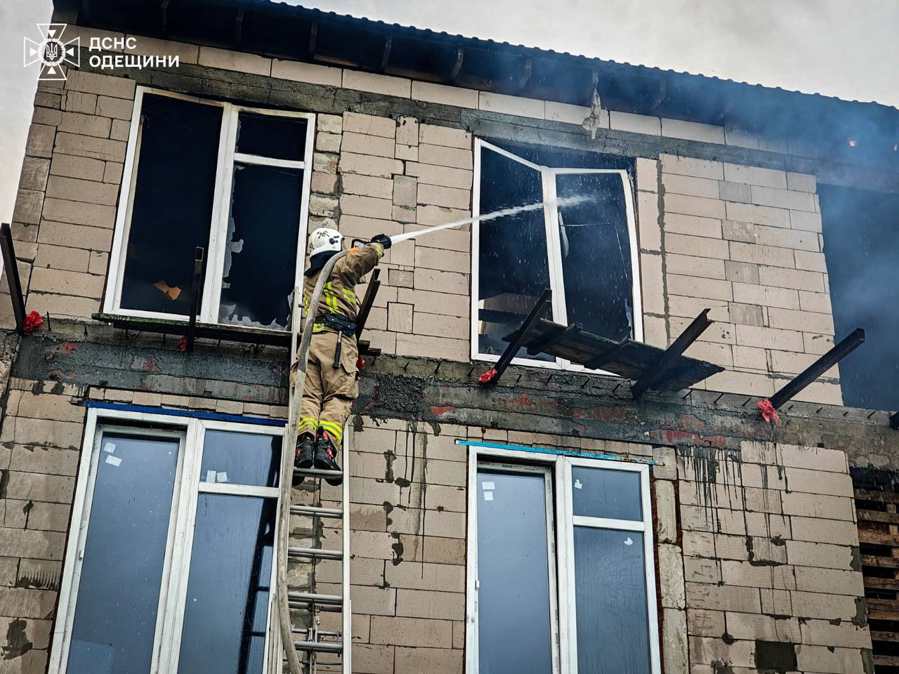 Рятувальник стоїть на висувній драбині та спрямовує струмінь води у вибите вікно другого поверху недобудованого будинку з газоблоків. З будівлі йде дим, а у верхньому лівому кутку розміщено логотип ДСНС Одещини.