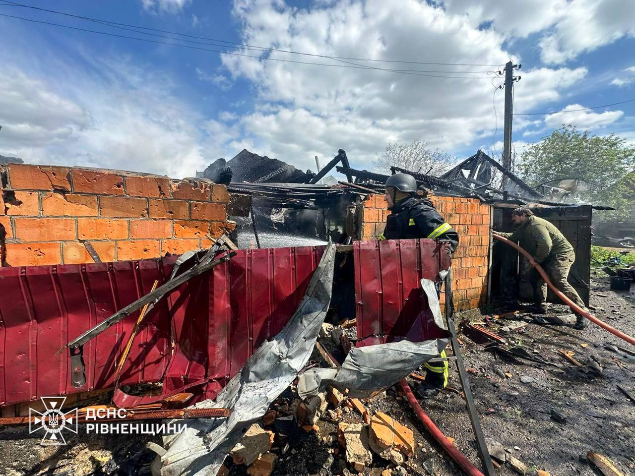 Рятувальник у формі та шоломі разом із чоловіком у цивільному одязі ліквідовують наслідки пожежі на території зруйнованої цегляної споруди. Навколо видно обвуглені конструкції, деформований металопрофіль та уламки цегли, а з лівого нижнього кутка б'є струмінь води. У лівому кутку розміщено логотип ДСНС Рівненщини.