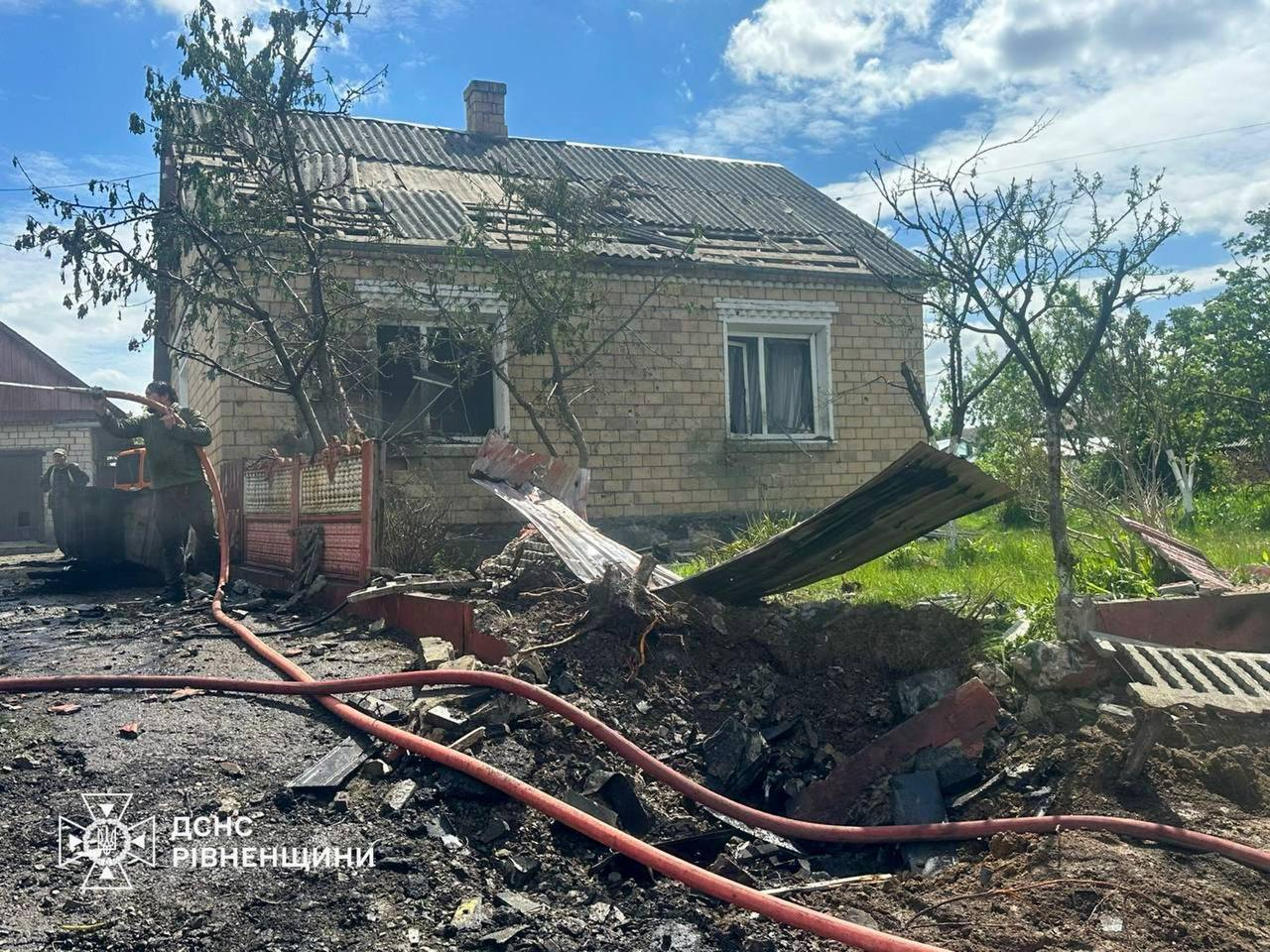 Наслідки ворожого обстрілу чи вибуху на подвір'ї приватного цегляного будинку. На передньому плані видно вирву в землі, розкидані листи шиферу, повалений паркан та пожежні рукави. Покрівля та вікна будинку частково пошкоджені, а ліворуч двоє чоловіків ліквідовують наслідки. У лівому кутку розміщено логотип ДСНС Рівненщини.