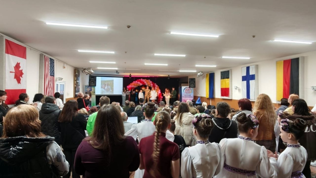Глядачі в залі, серед яких дівчата в українських вишиванках, спостерігають за виступом на сцені з великим екраном та аркою з повітряних кульок. На стінах приміщення розвішані прапори різних держав, зокрема Канади, США, України, Румунії, Фінляндії та Німеччини.