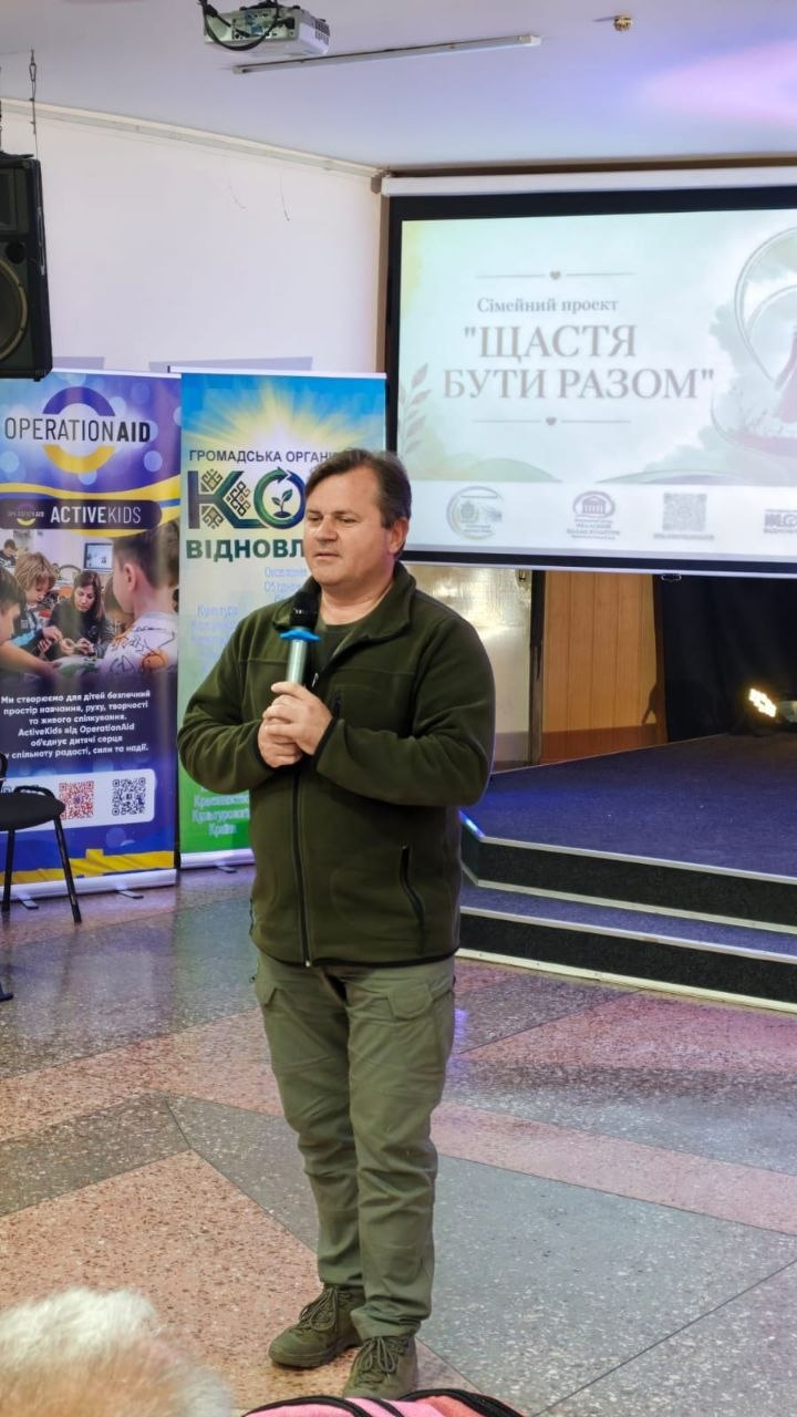 Чоловік у зеленій флісовій кофті та тактичних штанах виступає з мікрофоном у залі біля сцени. На задньому плані видно великий екран із написом «Сімейний проєкт 