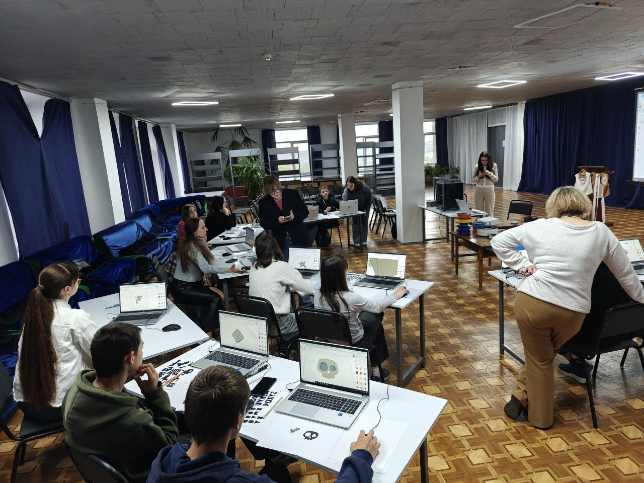 Люди працюють за ноутбуками у просторій залі з паркетною підлогою під час навчального заходу або комп'ютерного майстер-класу. Столи з технікою розставлені півколом, на екранах лептопів видно графічні моделі, а організатори консультують учасників.