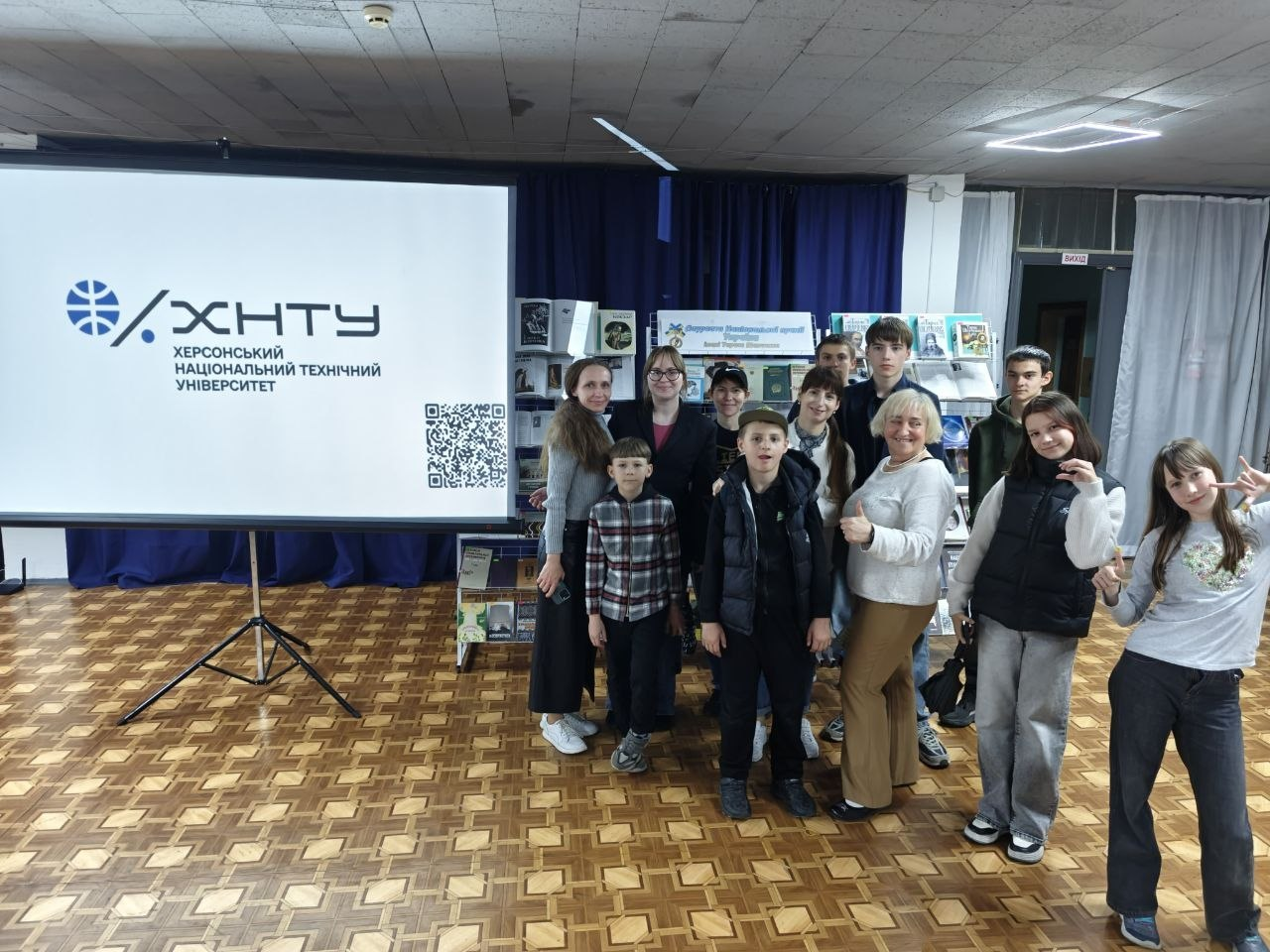 Група дітей та дорослих позує для фото в актовій залі або аудиторії. Ліворуч на штативі встановлено великий білий екран із логотипом «ХНТУ Херсонський національний технічний університет» та QR-кодом, а на задньому плані видно виставку книг.