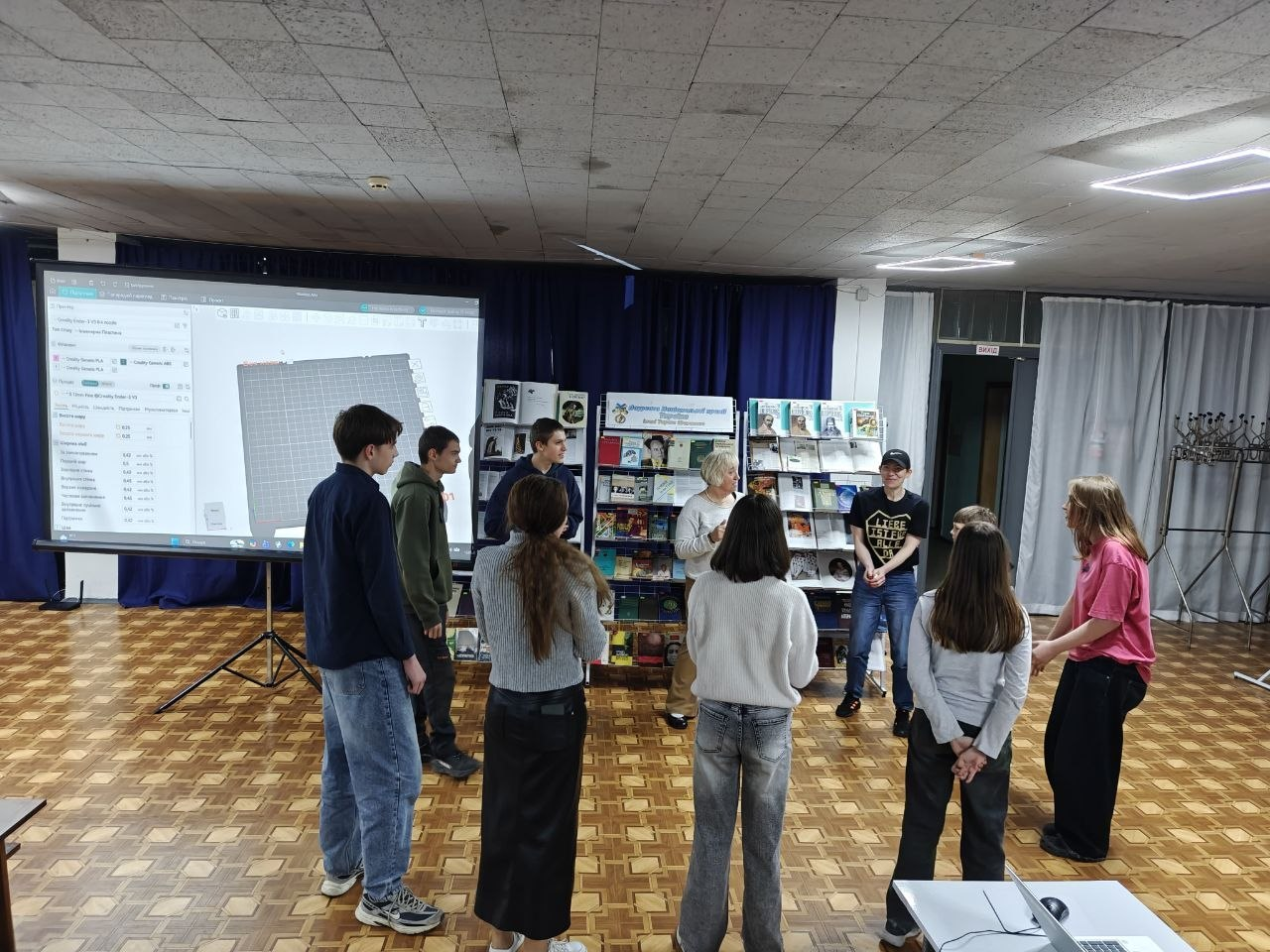 Група підлітків та дорослих стоїть колом в актовій залі, обговорюючи презентацію. Ліворуч на великому проекційному екрані відкрито інтерфейс програми для 3D-моделювання, а на задньому плані розташовані стенди з книжковою виставкою.