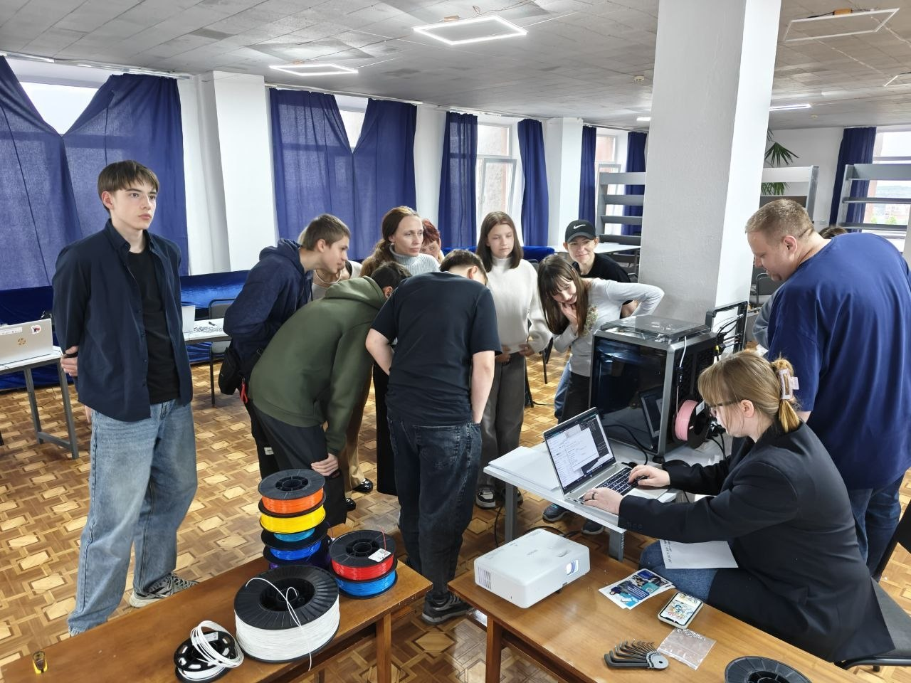 Група підлітків та дорослих зібралася навколо столу з обладнанням для 3D-друку в аудиторії. Жінка працює за ноутбуком, підключеним до 3D-принтера, а поруч на столі лежать котушки з кольоровим пластиком (філаментом).