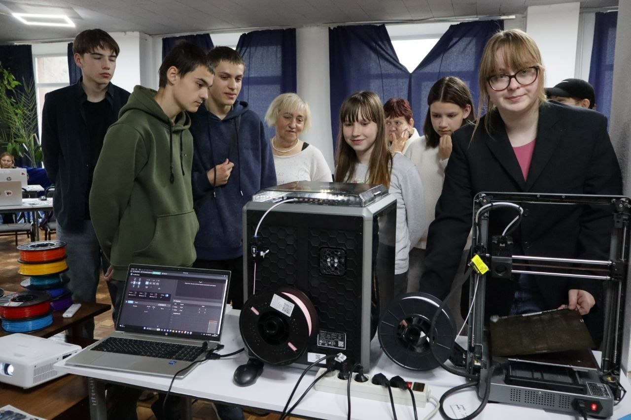 Група підлітків та дорослих спостерігає за роботою обладнання для 3D-друку в навчальній аудиторії. На передньому плані на столі стоять два 3D-принтери, відкритий ноутбук із програмою для моделювання та котушки з пластиком, а поруч стоїть дівчина в окулярах і піджаку.