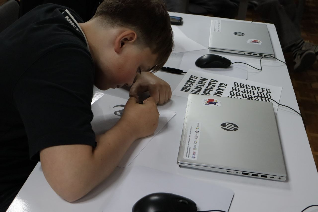 Хлопчик сидить за білим столом і зосереджено малює або пише на аркуші паперу. Поруч із ним на столі стоять два закриті сріблясті ноутбуки HP, лежать комп'ютерні мишки та роздруковані зразки латинського алфавіту в різних графічних стилях.