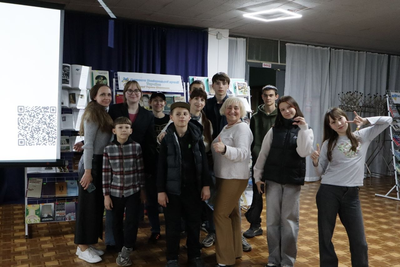 Група дітей та дорослих стоїть разом та позує для спільного фото в актовій залі чи аудиторії. Ліворуч видно частину великого білого екрана з QR-кодом, а на задньому плані розташовані стенди з книжковою виставкою під синьою завісою.