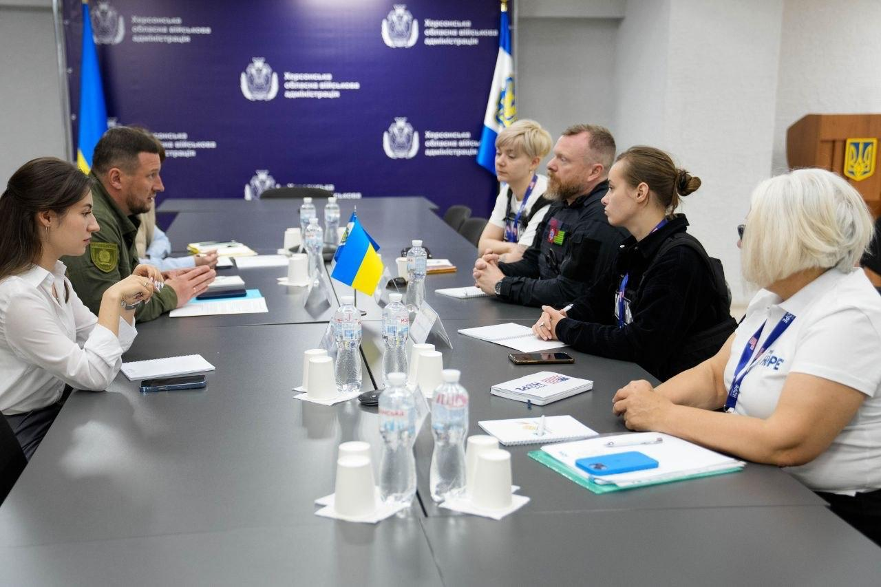 Офіційна зустріч двох делегацій за довгим сірим столом у конференц-залі. На столі розміщені пляшки з водою, паперові стаканчики, блокноти та маленький прапор України, а на синьому банері позаду видно емблеми та написи.